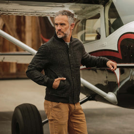 fit model wearing The Bomber Jacket in Charcoal Wool leaning against an airplane 